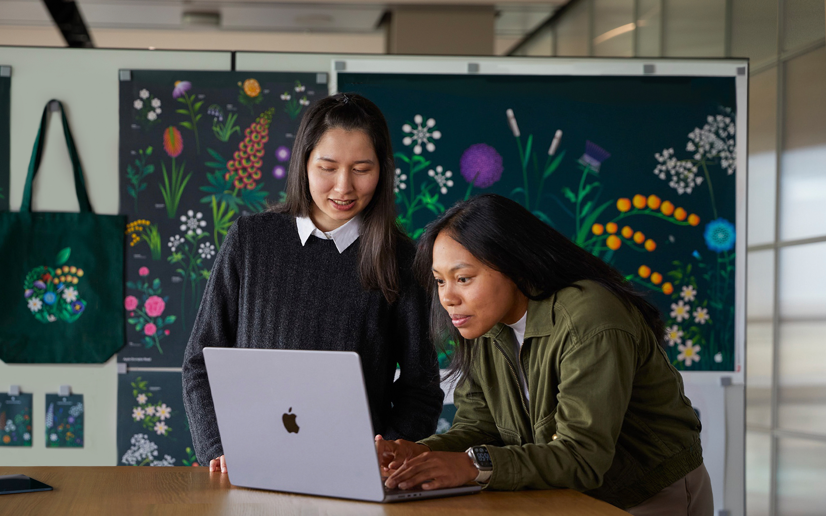 Twee teamleden van Apple Marketing die samenwerken op een MacBook. Ze zitten voor posters die hun team heeft ontworpen.