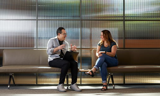 Dan sitting on an office bench with his MacBook on his lap and speaking to a colleague