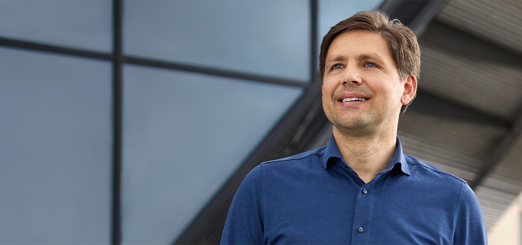 Greg a szabadban áll egy ablaktáblás fal előtt, és mosolyogva balra néz.