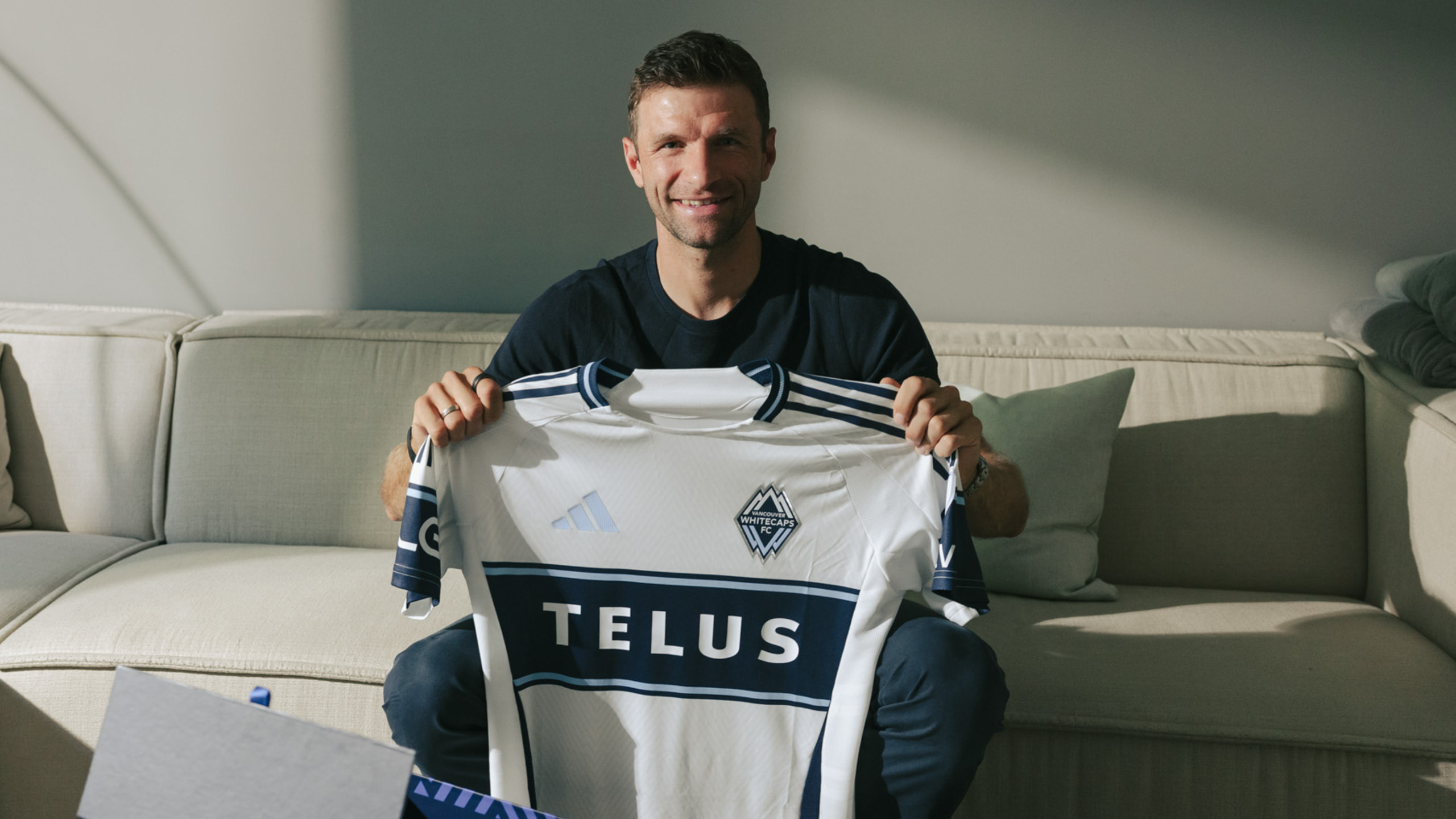 Thomas Müller, assis sur un canapé, pose avec un maillot du Vancouver Whitecaps FC.
