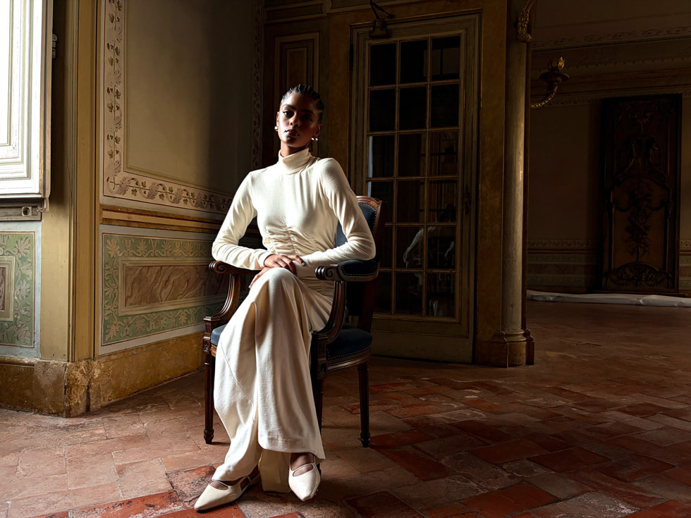 An editorial-style photo of a person wearing all white sitting on a chair inside a dimly lit room taken on iPhone Air.