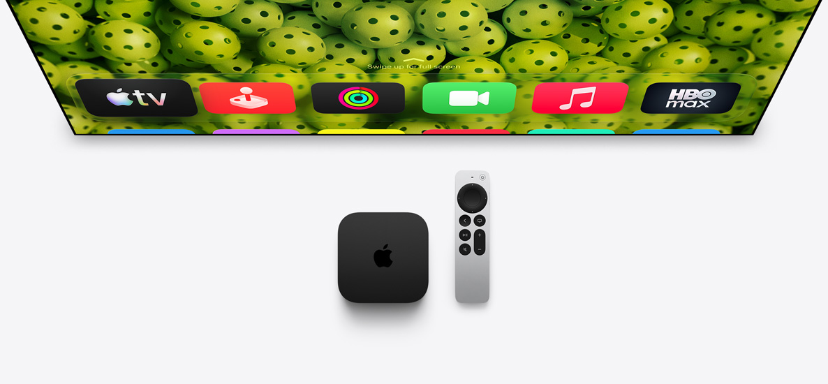 Apple TV 4K i pilot Apple TV Remote umieszczone pod ekranem telewizora, na którym widać ekran początkowy z tłem z zielonych piłek do pickleballa i z rzędem ikon aplikacji