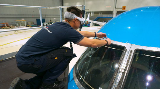 A technician in a plane hangar wears Apple Vision Pro while using a tool in their hands to adjust the plane's windshield