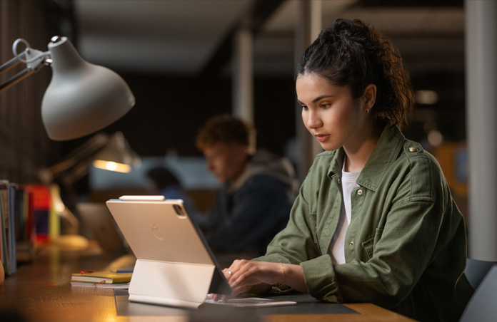 Una estudiante sentada en un escritorio en una sala de estudio escribe en un iPad con un Magic Keyboard para mostrar la versatilidad de los dispositivos Apple