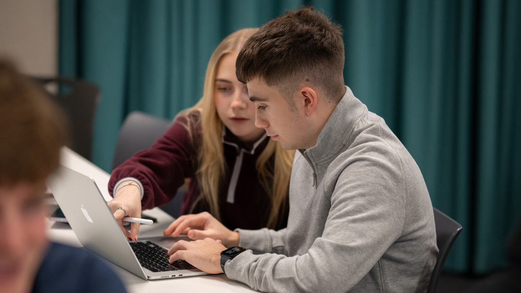 Compañeros de clase colaboran en un MacBook; ella señala la pantalla mientras él escribe
