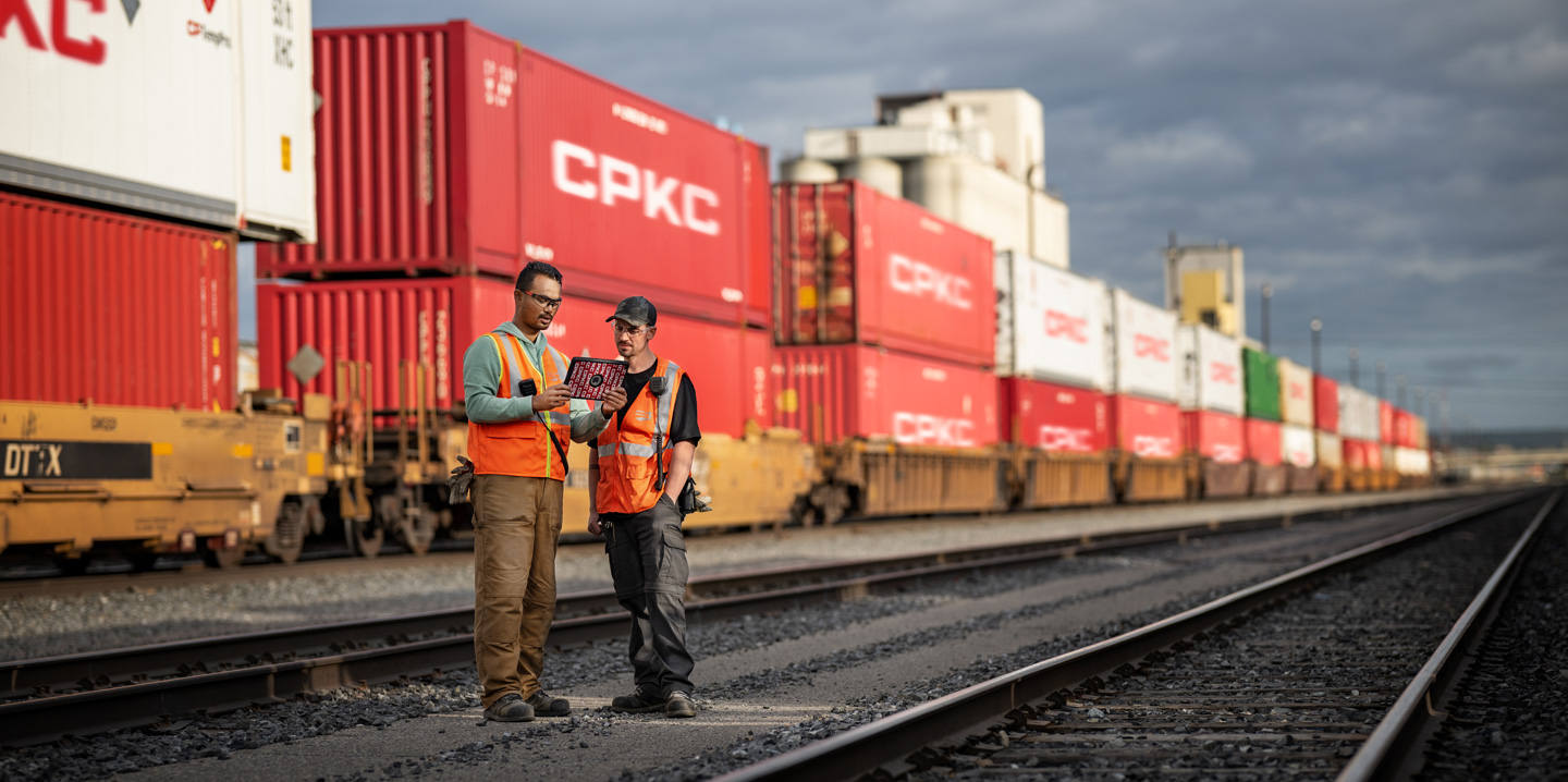 Twee spoorwegmedewerkers gebruiken een iPad terwijl ze op een rangeerterrein naast een stilstaande trein met zeecontainers staan