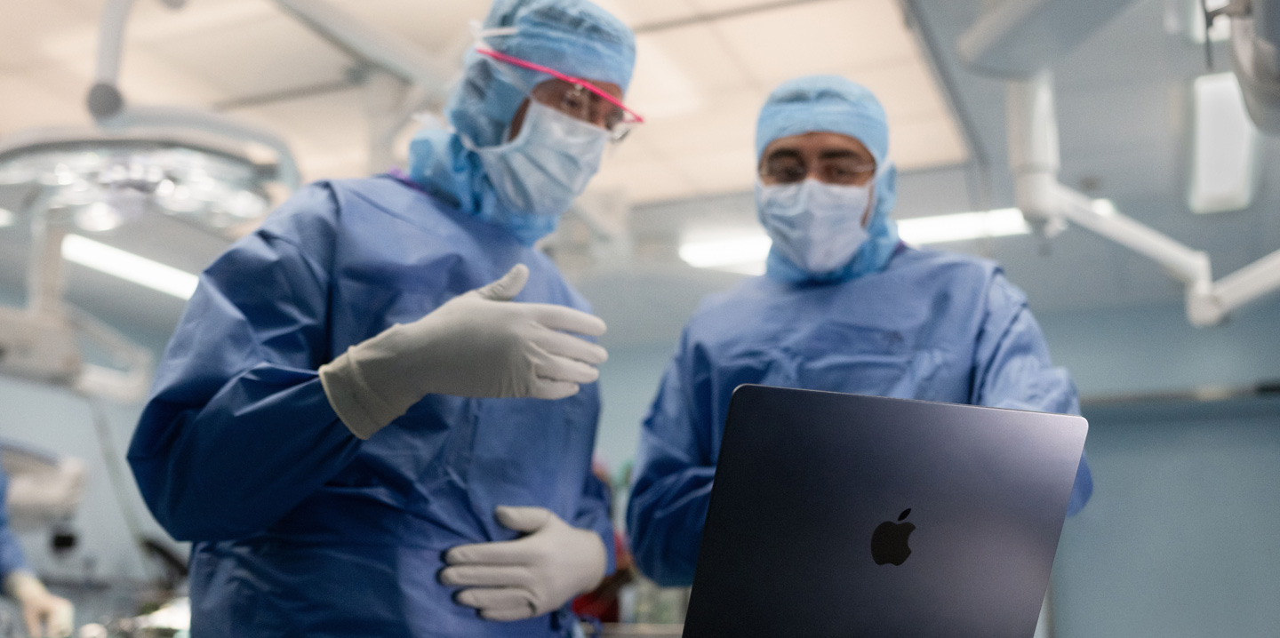 Deux chirurgiens dans une salle d’opération consultent un MacBook, l’un d’eux fait un geste de la main tandis que l’autre observe