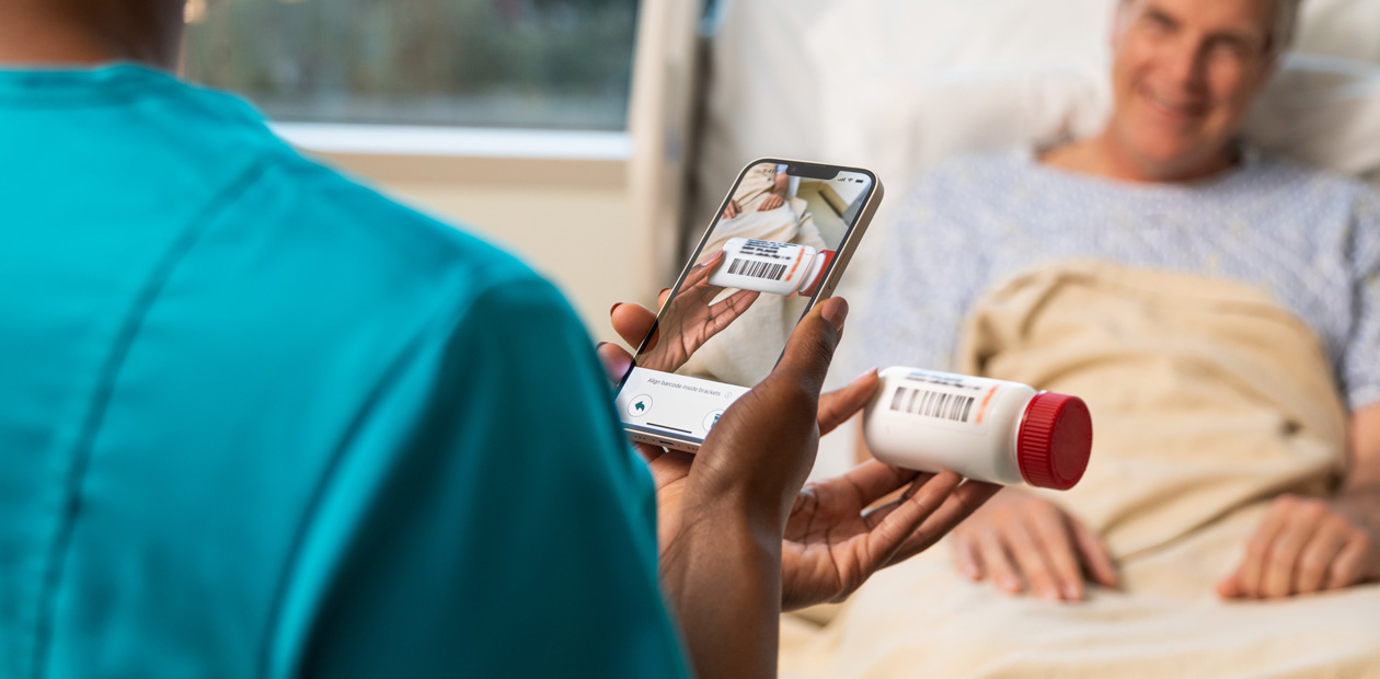 En sjuksköterska i blågröna sjukhuskläder som läser av en patients läkemedel med sin iPhone.